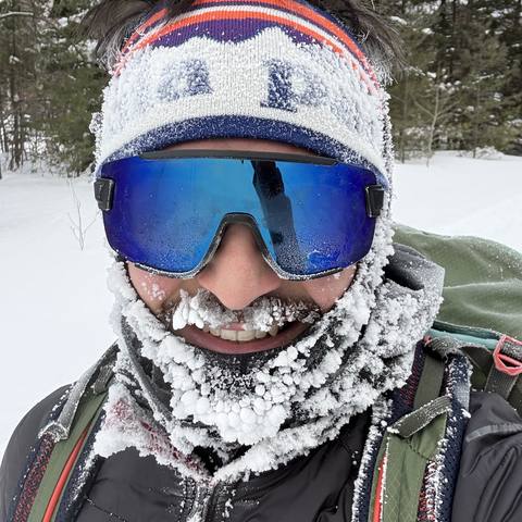 Félix Dubé outdoors in winter — ski goggles and a knit headband, beard and neck gaiter caked in frost, a pack over one shoulder and snowy conifers behind.